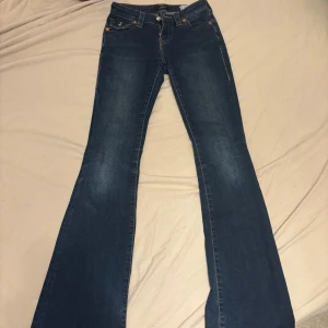 True Religion bootcut jeans blå - Snygga blå jeans från True Religion med klassiska kontrastsömmar och ikoniska bakfickor med lock och knappar. Modellen har bootcut-ben och låg midja, tillverkade i mjukt denimtyg. Perfekta för dig som gillar en tidlös och cool look.
