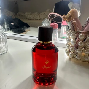 Alegria parfym röd flaska 100ml - Snygg parfym från Royal Platinum i en djup röd glasflaska med svart kork och gulddetaljer. Flaskan rymmer 100ml och har en elegant design med en liten ros och texten Alegria på framsidan. Perfekt för dig som gillar lyxiga och stilrena dofter.