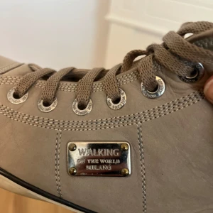 Beige sneakers från Walking Milano - Snygga beige sneakers från Walking Milano med högt skaft och rund tå. Skorna har snörning med metallöglor och en cool metallplatta med logga på sidan. Tillverkade i skinn med detaljerade sömmar och platt sula. Perfekt för dig som gillar stilrena och neutrala färger.