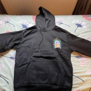 Svart hoodie Casablanca Avenida - Svart hoodie från Casablanca med stor färgglad print på ryggen och mindre tryck på bröstet. Snygg huva med snörning och klassisk känguruficka framtill. Perfekt för dig som gillar streetwear med unik design.