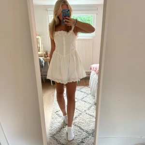 Vit kort klänning med spetsdetaljer - Supersöt vit kortklänning med broderade spetsdetaljer längs axelband och fåll. Klänningen har knappar framtill, markerad midja och volangig kjol. Perfekt för sommarens alla tillfällen och ger en romantisk vibe. Tillverkad i bomull för en luftig känsla.