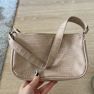 Beige axelväska i crocopräglat skinn - Snygg beige axelväska med crocopräglat mönster och guldfärgade detaljer. Väskan har en bred axelrem och stängs med dragkedja upptill. Insidan har ett mindre fack med dragkedja för småsaker. Perfekt storlek för det viktigaste.