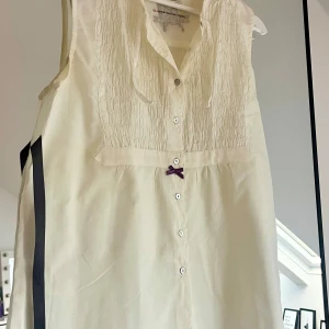 Ärmlös blus från Odd Molly - Supersöt vit blus från Odd Molly med smockad överdel, knappar hela vägen framtill och en liten lila rosett som detalj. Blusen är ärmlös och har en mjuk krage med knytband. Grått sidenband att knyta baktill. Fjäril i backen. Storlek 2 (M). Väl använd men ser fräsch ut. 