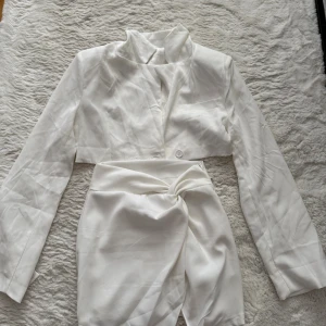 Vit kort set med knutdetalj - Stilren vit kort set med lång ärm och snygg knutdetalj framtill i midjan. Klänningen har en hög krage och är tillverkad i ett glansigt material som ger en lyxig känsla. Perfekt för dig som vill sticka ut med en modern och chic look.