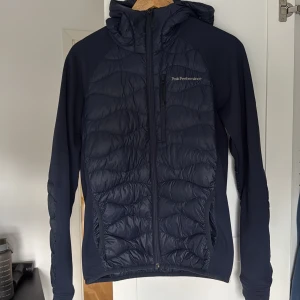 Mörkblå pufferjacka Peak Performance - Snygg mörkblå pufferjacka från Peak Performance med huva och hel dragkedja. Jackan har quiltade partier framtill, baktill och på ärmarna, samt släta sidor. Perfekt för kyliga dagar och enkel att matcha med olika outfits.