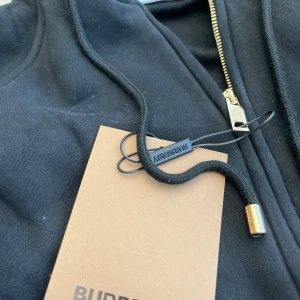 Svart hoodie från Burberry med dragkedja - Svart hoodie från Burberry med klassisk rutmönstrad huva, guldfärgad dragkedja och snörning. Tröjan har två fickor fram och ribbade muddar. Perfekt för dig som vill ha en stilren och lyxig look med ikoniska Burberry-detaljer.