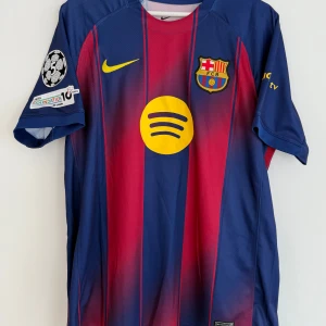 FC Barcelona hemmatröja 25/26 - Säljer en officiell FC Barcelona matchtröja från Nike med Lamine Yamal och nummer 10 på ryggen. Tröjan har klassiska blå och röda vertikala ränder, gula detaljer och klubbmärke på bröstet. Materialet är lätt och andas, perfekt för fotboll eller häng. Spotify-logga på framsidan.
