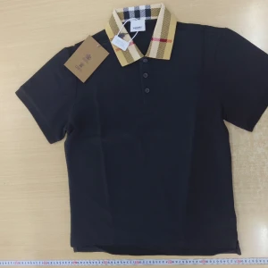 Svart Burberry t-shirt med rutig krage - Svart t-shirt från Burberry med klassisk rutig krage i beige, svart och rött. Modellen har korta ärmar och tre knappar framtill. Snygg och stilren design som lyfter din outfit med lyxig känsla. Materialet är mjuk bomull för skön komfort.