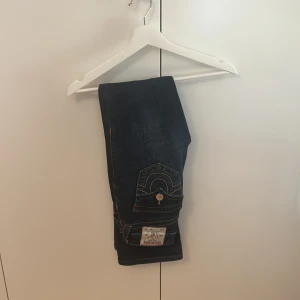 True Religion Becca Mid Rise Bootcut Jeans - Mörkblå jeans från True Religion, modell Becca Mid Rise Bootcut. Snygga kontrastsömmar och ikoniska fickdetaljer med knappar bak. Klassisk bootcut-passform och tillverkade i slitstarkt denim. Perfekta för dig som gillar en tidlös och cool look. Dom är i storlek 25 och är lite uppsydda i benen. Har använt 2 gånger. 