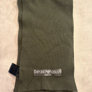 Olivgrön halsduk Emporio Armani - Snygg olivgrön halsduk från Emporio Armani Parfums med diskret logotyp framtill. Halsduken är lång och smal, stickad i mjukt syntetmaterial som känns skönt mot huden. Perfekt för att ge din outfit en clean och stilren touch.