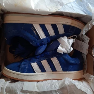 Adidas Campus blå sneakers - Snygga Adidas Campus sneakers i blå mocka med klassiska vita ränder på sidorna och vit sula. Skorna har rund tå, snörning och en retro vibe som passar perfekt till streetwear. Adidas-logga och Campus-tryck på sidan ger en ikonisk look.