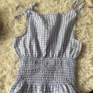 Blårutig topp/linne  - Supersöt blårutig topp i storlek M.  Linnet har smockad midja, tunna axelband med knyt och ärmlös design. Perfekt för varma dagar med sitt lätta material och fräscha färg. Passar dig som gillar en chill och trendig look. Ny endast testad