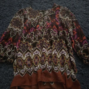 Mönstrad blus från Lindex i lila och rött - Säljer en blus från Lindex med färgstarkt mönster i lila, rött, svart och beige. Blusen har långa ärmar, knappar framtill och luftig passform. Perfekt för dig som gillar unika prints och sköna vibes.