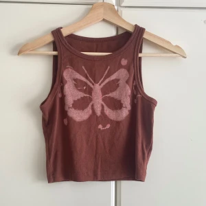 Brun croppad linnetopp med fjäril - Säljer en brun croppad linnetopp med ett stort rosa fjärilstryck framtill. Toppen är ärmlös och har en enkel, rak passform. Perfekt för varma dagar och ger en cool vibe till din outfit.