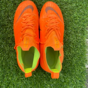 Nike Flyknit orange fotbollsskor - Säljer ett par grymt snygga fotbollsskor från Nike i en riktigt skrikig orange färg med svarta detaljer och Nike-logga. Skorna är tillverkade i lätt och smidig flyknit med snörning och har gröna innersulor. Perfekta för dig som vill sticka ut på planen!