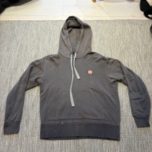 Grå hoodie från Acne Studios - Säljer en grå hoodie från Acne Studios med klassisk face patch på bröstet. Tröjan har huva med snörning, känguruficka och långa ärmar. Bra skick förutom ett litet hål vid snörningen. Annars har den från början en ”distressed” look. 