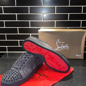 Christian Louboutin grå mocka sneakers - Snygga grå sneakers från Christian Louboutin i mocka med ikoniska röda sulor. Skorna har rund tå, snörning och coola nitar på tån för en edgy vibe. Perfekta för dig som vill sticka ut med lyxig streetstyle.