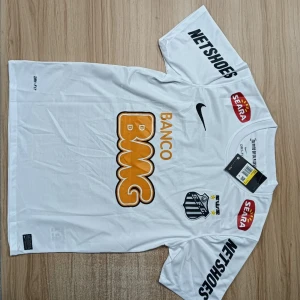 Santos Neymar Jr 11 vit fotbollströja Nike - Säljer en officiell Santos FC fotbollströja med Neymar Jr och nummer 11 på ryggen. Tröjan är vit med svarta och orangea tryck, korta ärmar och rund hals. Materialet är lätt och andas, perfekt för match eller träning. Sponsorer som BMG, Netshoes och Seara syns tydligt.