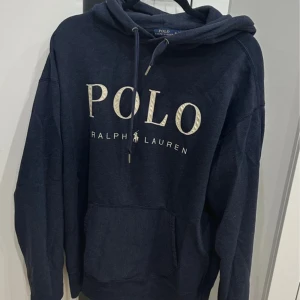 Mörkblå hoodie från Polo Ralph Lauren - Snygg mörkblå hoodie från Polo Ralph Lauren med stor logga framtill. Tröjan har huva med snörning och en klassisk känguruficka. Perfekt för en chill och avslappnad stil. Tillverkad i mjuk bomullsmix för extra komfort.