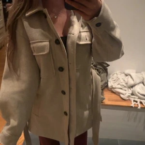 Beige overshirt med bälte och fickor - Snygg beige overshirt med stora bröstfickor, knappar framtill och ett matchande bälte i midjan. Jackan har en rak och loose passform med klassisk krage och långa ärmar. Perfekt för lager-på-lager och enkel att styla till jeans eller leggings.