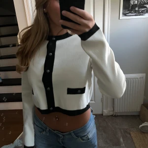 Vit och svart croppad kofta - Säljer en stilren croppad kofta i vitt med svarta detaljer och knappar framtill. Koftan har långa ärmar, svarta kanter vid ärmslut och nederkant samt dekorativa fickor framtill. Perfekt för dig som gillar en trendig och klassisk look.