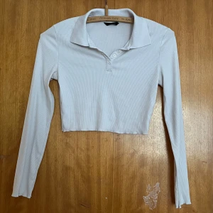 Vit croppad polotopp med knappar - Croppad polotopp i ljusvit färg med ribbad struktur. Toppen har långa ärmar, klassisk krage och fyra vita knappar framtill. OBS! Lite nopprig❤️