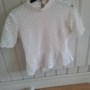 Vit spetsblus med volangkant - Superfin vit blus i spets med korta ärmar och volangkant nedtill. Blusen har hög hals med små bollar som detalj och knäppning i nacken. Perfekt för dig som gillar romantisk och söt stil. Den har en liten smink fläck vid halsen går särket bort i tvätten