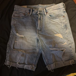 Blå slitna jeansshorts från H&M - Säljer ett par blå jeansshorts från H&M i regular fit med råa, fransiga kanter och slitna detaljer framtill. Shortsen har klassisk femficksdesign och är gjorda i denim med en cool tvättad look. Perfekt för dig som gillar streetstyle och avslappnad vibe. Den är helt ny