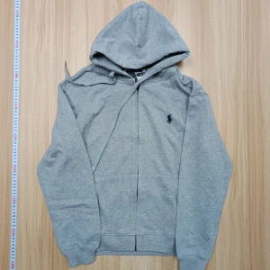 Grå hoodie från Polo Ralph Lauren M - Klassisk grå hoodie från Polo Ralph Lauren i mjuk bomull. Tröjan har huva med snörning, dragkedja framtill och den ikoniska broderade loggan på bröstet. Perfekt för en chill och stilren look.