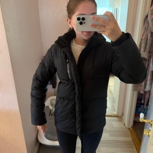 Svart pufferjacka från Calvin Klein - Säljer en svart pufferjacka från Calvin Klein Jeans med huva och dragkedja. Jackan har logga på ärmen och är vadderad för extra värme. Perfekt för kyliga dagar och har en clean, stilren look som funkar till det mesta.
