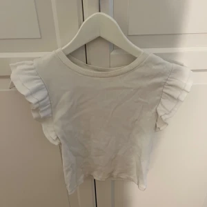 Vit ribbad topp med volangärmar - Supersöt vit ribbad topp med volangdetaljer på ärmarna. Toppen har rund halsringning och är tillverkad i ett mjukt bomullstyg. Perfekt för dig som gillar en feminin och fräsch stil med extra detaljer på axlarna. Säljer då den inte passar mig längre.💗