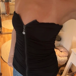 Svart bandeau tubtopp med dragkedja - Säljer en svart bandeau tubtopp från Guess med snygg dragkedja framtill och rynkade detaljer längs hela toppen. Storlek står ej men är syretchig av sig så skulle passa från XS till S/M