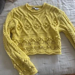 Gul stickad tröja från Zara - Säljer en superfin gul stickad tröja från Zara i storlek S. Tröjan har ett unikt mönster med flätor och bollar, samt en croppad passform. Perfekt för att liva upp garderoben med lite färg! Rund halsringning och långa ärmar.