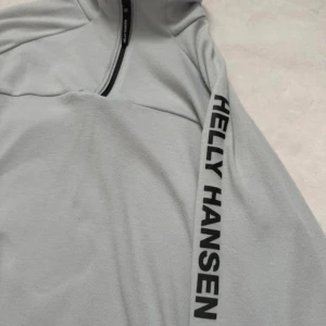 Ljusgrå Helly Hansen fleece half zip - Ljusgrå fleece-tröja från Helly Hansen med svart logga längs ärmen och HP Hydropower-detalj. Tröjan har half zip framtill, hög krage och är långärmad. Perfekt för kyliga dagar och outdoor vibes.