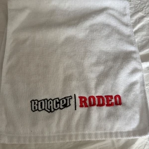 Bolaget Rodeo 2024 handduk - Vit handduk med broderad Bolaget Rodeo-logga i svart och rött. Tjockt, mjukt tyg. Kastad av Oliwer i från konserten i Luleå. Har nå små fläckar som lätt går bort i tvätten om man skulle vilja att jag gör de.