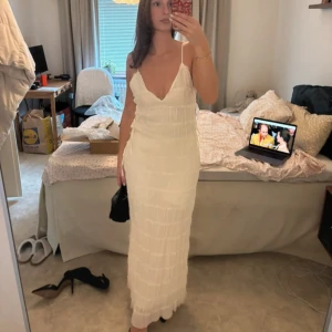 Vit långklänning Positano Style - Superfin vit långklänning i Positano-stil med smala axelband, v-ringning och knyt i sidorna. Klänningen har ett plisserat tyg med horisontella sömmar som ger en unik struktur. Perfekt för sommarens alla tillfällen!