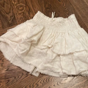 Vit volangkjol från Zara, stl 140 - Supersöt vit kortkjol från Zara med volanger och smockad midja med knytband. Kjolen är i tunn, luftig bomull. Storlek 140 cm