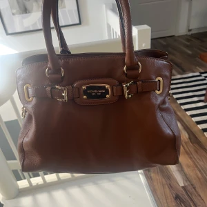 Brun handväska från Michael Kors - Snygg brun handväska från Michael Kors i skinn med guldfärgade metalldetaljer och spännen framtill. Väskan har dubbla handtag, avtagbar axelrem och flera fack inuti med logotypmönstrat foder. Perfekt för dig som vill ha både stil och funktion.