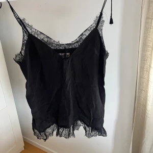 Svart linnetopp med spetsdetaljer - Säljer en svart linnetopp med tunna axelband och v-ringning. Toppen har snygga spetsdetaljer längs halsringning och nederkant som ger en extra touch. Perfekt att styla med jeans eller kjol för en trendig look.