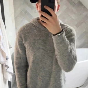 Grå fluffy stickad hoodie med huva - Mysig grå hoodie i stickat material med huva och lång ärm. Tröjan har en fluffig och avslappnad look, perfekt för kyliga dagar. Passformen är normal och den neutrala färgen gör den enkel att matcha med jeans eller andra plagg.