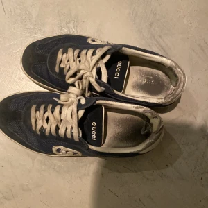 Mörkblå sneakers från Gucci - Snygga mörkblå sneakers från Gucci med klassisk GG-logga på sidan. Skorna är ganska trasiga i hällen Skorna har vita snören, vit sula och detaljer i mocka och textil. Insidan är svart med Gucci-tryck. Perfekta för dig som gillar exklusiva och stilrena sneakers. 