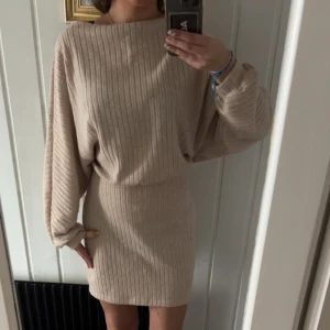 Beige ribbad stickad kortklänning - Supermysig beige stickad klänning med ribbat mönster och ballongärmar. Klänningen är kort och har ett knytband i midjan som ger en snygg siluett. Perfekt för dig som gillar en chill men ändå stilren look.