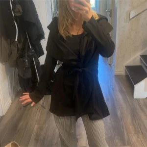 Svart omlott trenchcoat från L.B.C. - Svart trenchcoat med snygg omlottmodell och bälte i midjan från L.B.C. Jackan har en stor huva, långa ärmar och en loose passform. Perfekt för dig som vill ha en stilren och enkel jacka med coola detaljer.