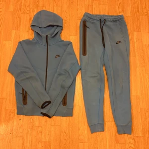 Blå Nike tech fleece tracksuit - Säljer ett par blå mjukisbyxor från Nike med svarta dragkedjor på fickorna och snörning i midjan. Byxorna har en slim passform och muddar vid bensluten. Perfekta för chill eller träning, riktigt sköna och stilrena.