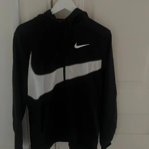 Svart Nike hoodie med vit swoosh - Svart hoodie från Nike med stor vit swoosh-logga tvärs över bröstet och dragkedja framtill. Tröjan har huva med snörning och långa ärmar. Perfekt för en sportig och avslappnad stil.