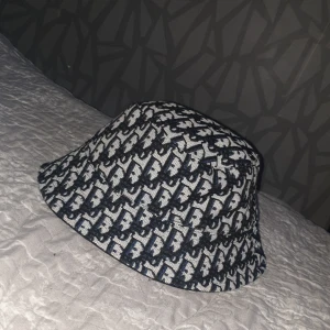 Mönstrad bucket hat från Off Work - Bucket hat med coolt svart och vitt mönster från Off Work. Hatten har en klassisk rund form och är tillverkad i ett slitstarkt tyg, troligen bomull. Insidan är helsvart och har en snygg etikett med märket. Perfekt för att lyfta din streetwear-stil.