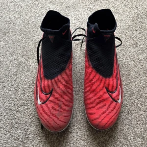 Nike röda och svarta fotbollsskor - Snygga fotbollsskor från Nike i rött och svart med coolt mönster och stora vita swoosh-loggor. Skorna har snörning, högre strumpliknande skaft och dobbar för gräsplan. Ovandelen är i syntetmaterial och designen är riktigt modern.    I just have the boots for these. No bag