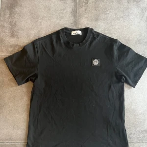 Svart Stone Island t-shirt i bomull - Svart t-shirt från Stone Island med klassisk rund hals och korta ärmar. På bröstet sitter den ikoniska Stone Island-loggan i svart och vitt. Tillverkad i mjuk bomull som känns skön mot huden. Perfekt för dig som gillar stilrena och enkla plagg med streetkänsla.