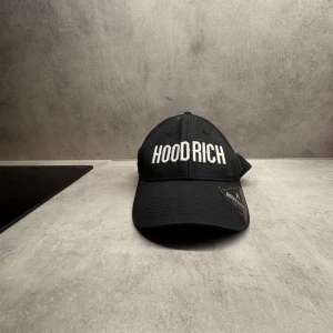 Svart keps från Hoodrich - Svart keps från Hoodrich med vit broderad logga framtill. Justerbar rem med metallspänne baktill för perfekt passform. Tillverkad i 100% bomull och har autenticitetsmärke på skärmen. Snygg och enkel design som passar till streetwear.
