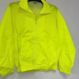 Neongul vindjacka - Säljer en neongul vindjacka med dragkedja framtill. Jackan har en hög krage och elastiska muddar vid ärmslut och nederkant. Perfekt för att synas i alla väder!